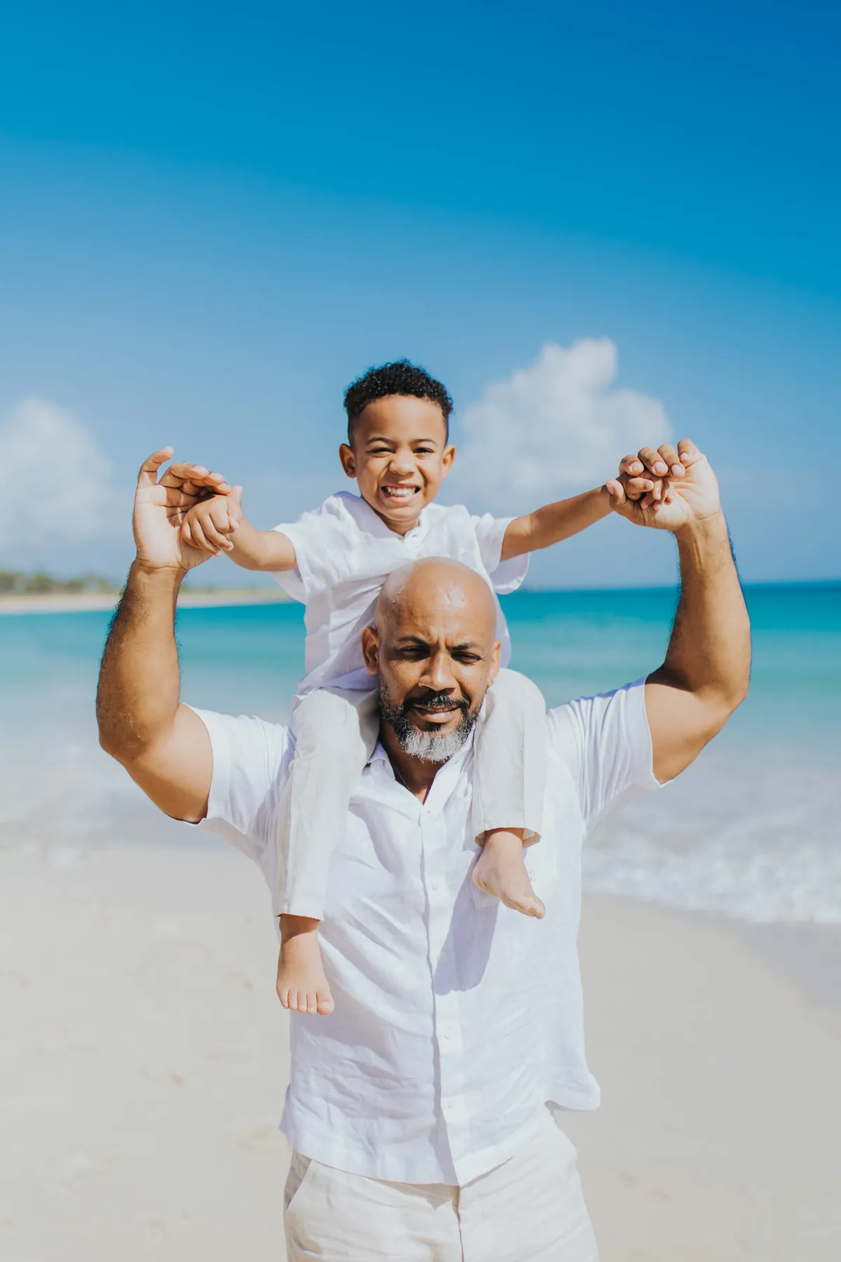 Boy on his dad's shoulders with arms up, turquoise sea behind