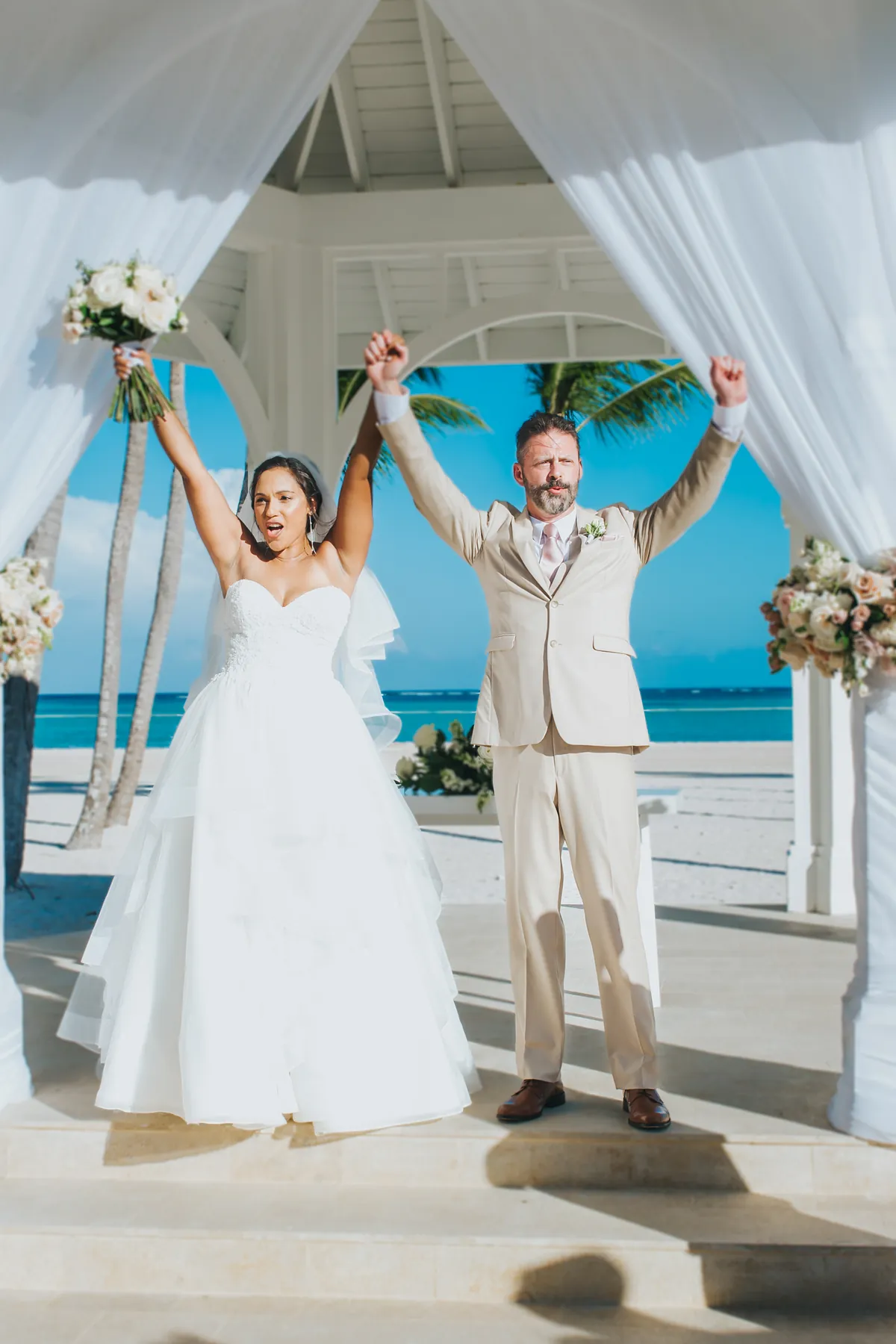 Newlyweds celebrating with arms raised under a beach gazebo in Punta Cana