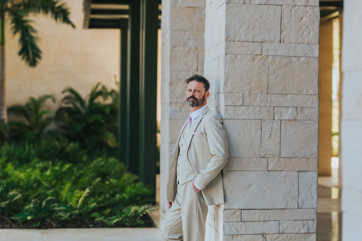 Groom in a beige suit leaning on a stone column